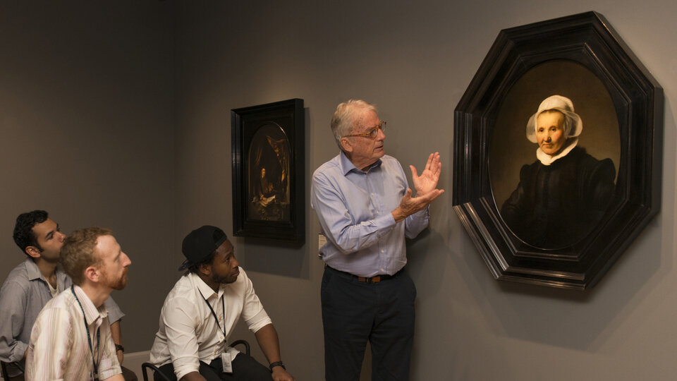 A man stands alongside a painting of a seated elderly woman wearing a white headpiece and black top. The standing man gestures to the painting while students listen intently, seated on stools nearby
