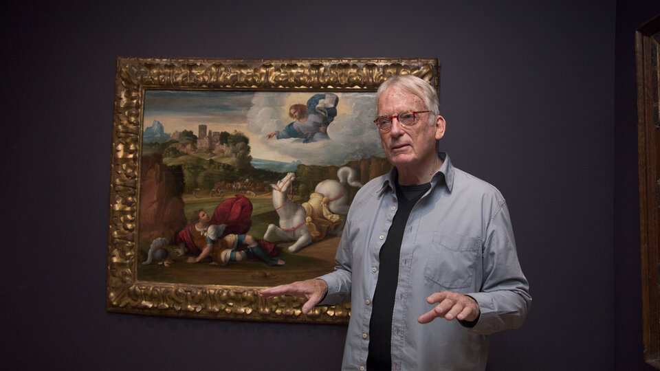 A man stands with his back to a painting of a scene that includes a man who has fallen off his horse, both of which are looking up at an angel in the sky. The man in the foreground gestures and appears to be talking to someone who is not in view of the camera