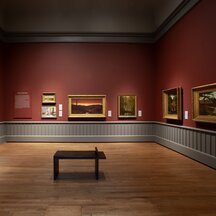 A view of an installation in a gallery with wooden floors and dark-red walls. Paintings hang alongside a single panel of text. A doorway is visible at far left, and a bench stands in the middle of the room.