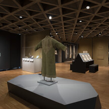 A gallery installation. Objects are presented on walls, on platforms, and in vitrines throughout the space. These include a green robe, displayed on an armature in the center foreground.