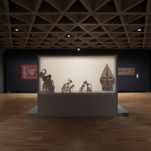 A gallery installation with shadow puppets displayed behind a screen, textiles hanging on walls, and other objects on platforms in the space.
