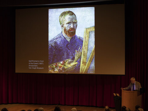 A darkened lecture hall with an image on the screen of a red headed man at an easel. At the podium a man stands