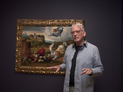 A man stands with his back to a painting of a scene that includes a man who has fallen off his horse, both of which are looking up at an angel in the sky. The man in the foreground gestures and appears to be talking to someone who is not in view of the camera