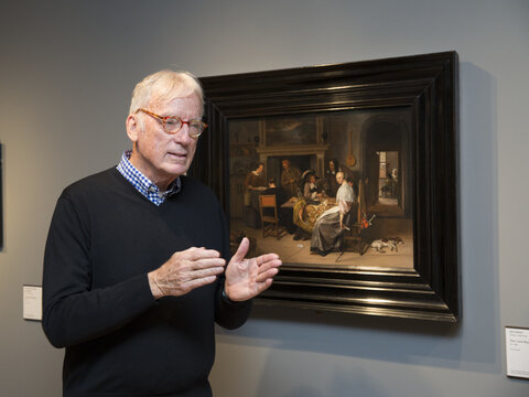 A man stands alongside a painting of people sitting in an interior space. The man gestures as though talking to someone who is not in view of the camera