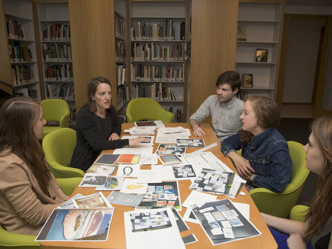Students and educator sit around a large table with papers scattered about. The group is situated in a library and are deep in discussion