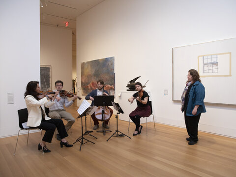 Four people playing string instruments while seated in a gallery. A fifth person stands nearby, watching them.