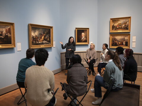 A group seated in a semicircle around an artwork that hangs on the wall. A standing figure gestures toward the artwork.