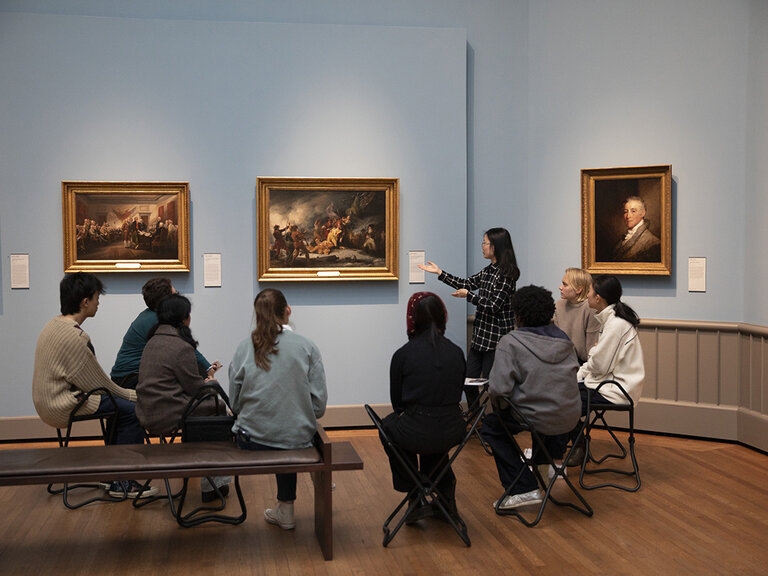 A group of people seated on stools and a bench in a gallery while viewing a work of art that hangs on the wall. Next to the artwork, one person stands and gestures.