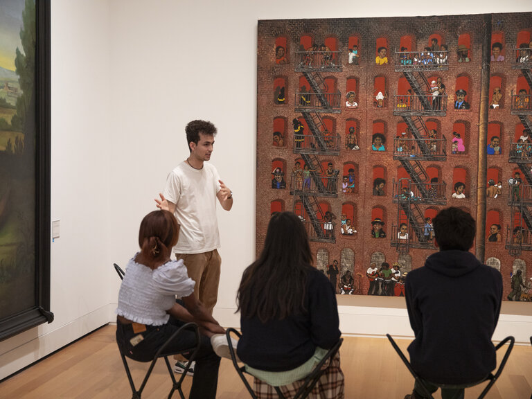 A group of people seated on stools and seen from behind as they view of work of art that hangs on the wall. Next to the painting, a person stands facing the group while gesturing.