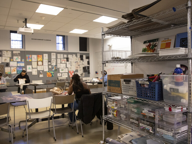 A classroom and/or studio space. In the right foreground stands a tall shelf with art supplies, books, and other materials. In the left background, three people sit at tables configured in a square or circle.