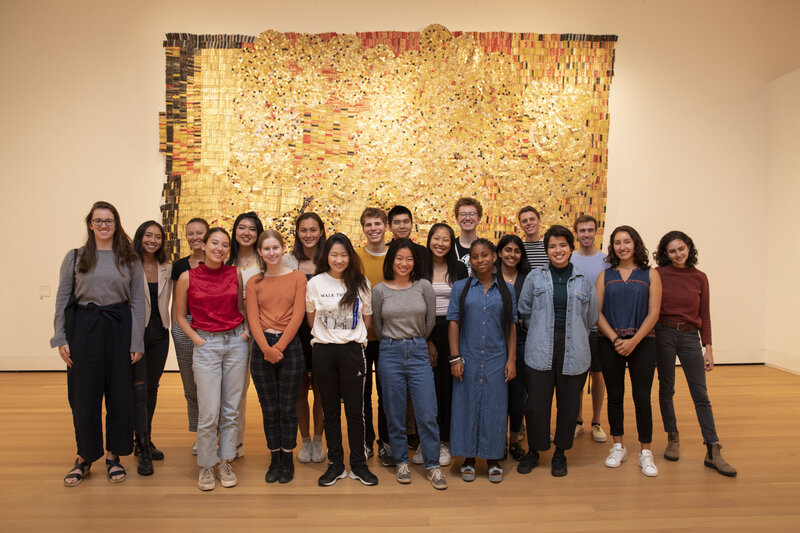 Twenty students in front of El Anatsui's golden sculpture made of aluminum bottle caps and copper wire.