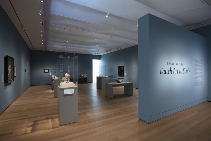 View of an exhibition with a title wall to the right that says "Thinking Small: Dutch Art to Scale"