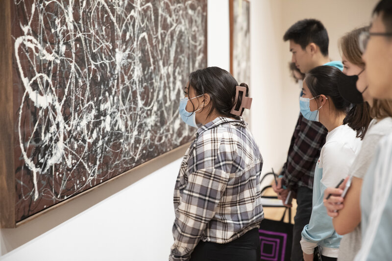 A large painting of paint splatters hangs on a white wall while students look closely at it