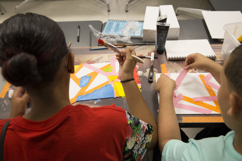 Teens seated at a table, with backs turned, engaged in the process of art making using colorful tape and paint on white paper