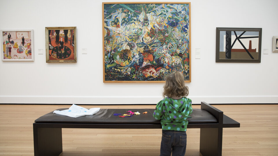 A child wearing a green top stands at a bench facing a wall of paintings. The child faces the artwork and has felt splayed out across the bench