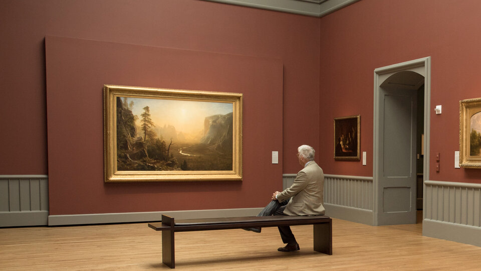 A man sits on a bench, with his hands clasped around his knee, looking at a large landscape painting hung on a maroon colored wall