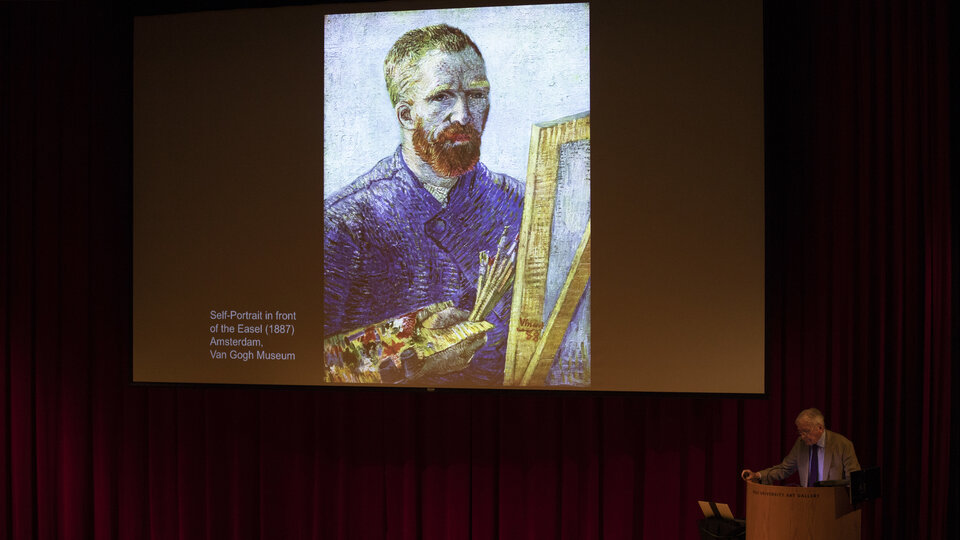 A darkened lecture hall with an image on the screen of a red headed man at an easel. At the podium a man stands