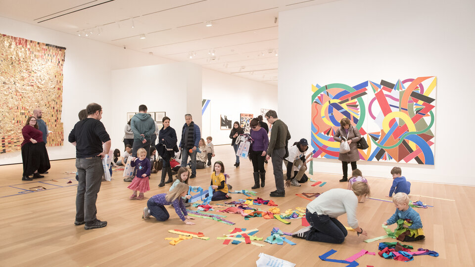 Families gather in a large exhibition space, engaging in activities with colorful ribbons on the floor