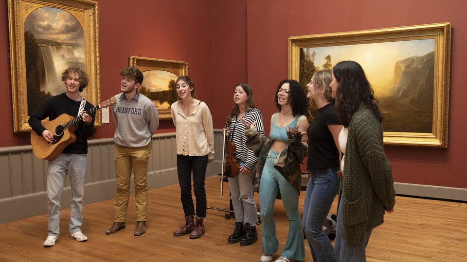 A group of students sing and play guitar in a red-walled gallery with landscape paintings on the walls behind them