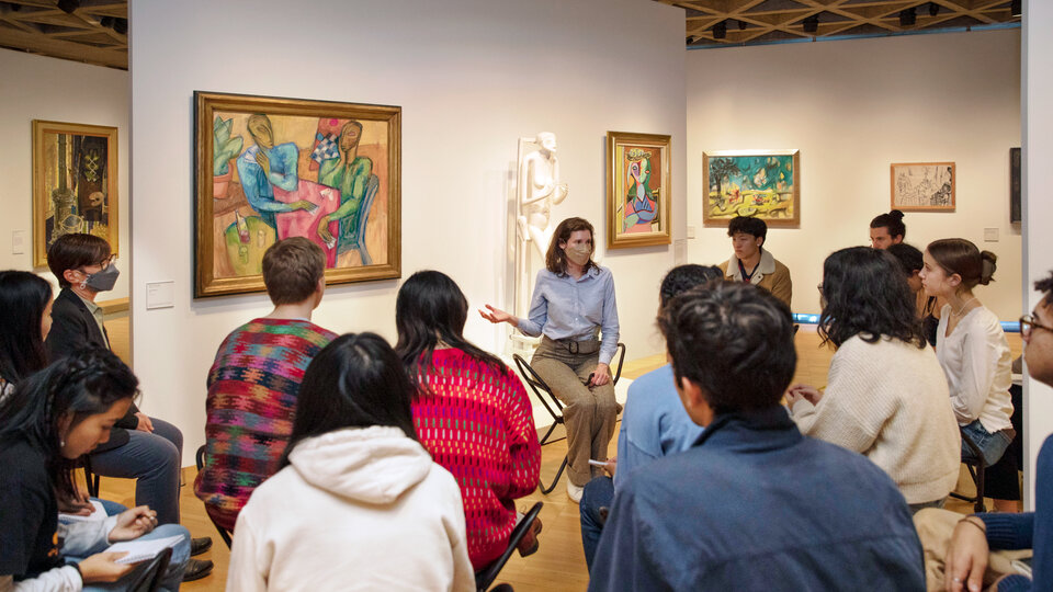 A group of young people seated in a gallery. They face a seated woman who wears a surgical mask and gestures with her right hand toward a painting that hangs on the wall next to her. The painting is colorful and abstract and shows two people playing cards across a table. Other artworks hang on the surrounding walls, and a sculpture stands on a base behind the seated woman.