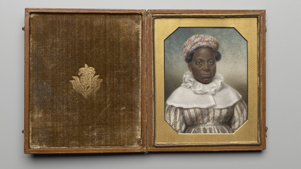 A painting of a Black woman from the waist up gazing out at the viewer housed in a hinged case. She wears a dress with vertical stripes and a striped headwrap that leaves her blackish-gray hair visible along the hairline. Over her shoulders and neck, she wears a lightweight, white garment with a high ruffled collar. 