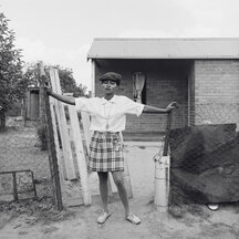 A black-and-white photograph of a woman standing outdoors between two sections of a chain-link fence. Holding her arms out in a T, with either hand grasping a fencepost, she addresses the camera with a confident gaze. The woman wears a dark pageboy hat and a white polo shirt tucked into a short, plaid skirt. In the background is a low brick building with a slanted roof.