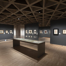 A gallery installation with gray walls, on which hang framed prints and drawings. At the center is a narrow, rectangular glass-covered display case with two, long unfolded lithographs on paper.