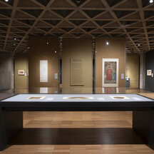A gallery installation with 3 artworks in a rectangular, covered display case in the foreground, and in the background, two paintings hang on tan walls with a text panel in between them.