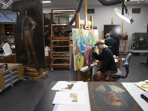 Conservator seated at an easel working on painting in conservation lab. Numerous other paintings of varying sizes on easels fill the space while a second conservator works on a large painting in the background. 