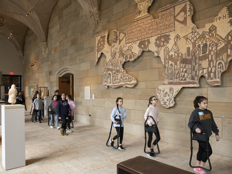 Visiting middle school students walking in a line through the sculpture gallery carrying portable chairs.