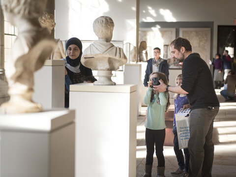 Man helping a young girl take photos of a marble sculpture, while a woman stands nearby looking at another work in the Ancient art gallery.