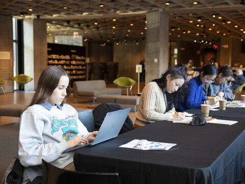 Female Yale student using her laptop that's sitting on a long table with a black tablecloth. Three other female students are sitting nearby and writing on pieces of paper.