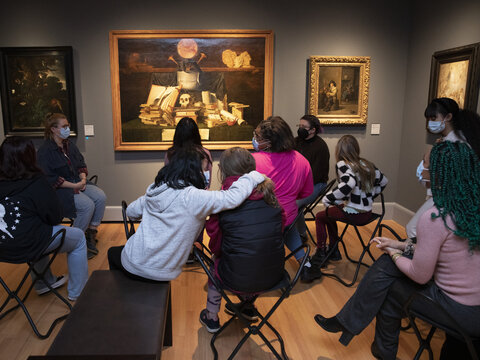 A group of people sit on stools in a gallery with their backs to the camera. They are looking at a painting