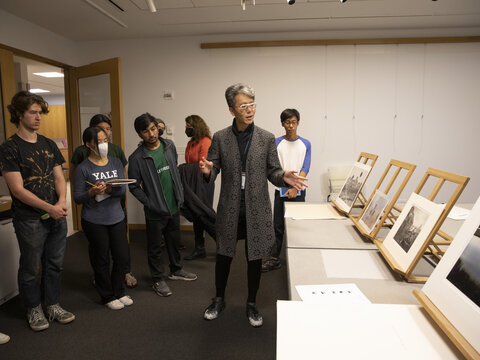 A man stands in the center of a room, gesturing to photographs on easels on a table to his left. A crowd of students stand around looking at the photos