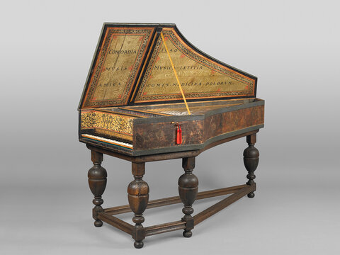 A keyboard instrument resembling a piano. Text and ornamentation are visible on the inside of its raised lid and on the surfaces above the keyboard itself. The instrument has elaborately carved legs.