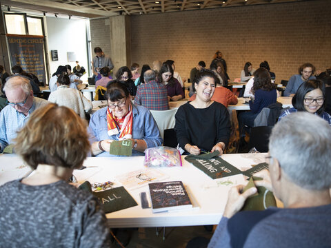 People seated at tables in a large room. Each appears to be sewing text into a piece of green fabric.