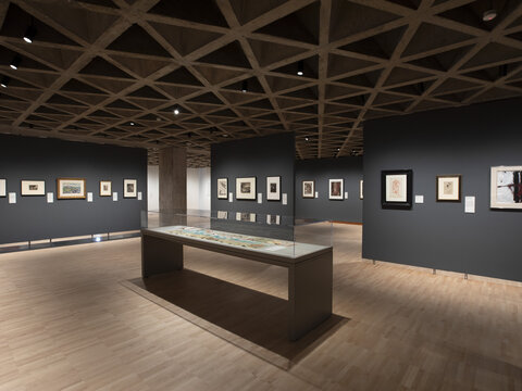 A gallery installation with gray walls, on which hang framed prints and drawings. At the center is a narrow, rectangular glass-covered display case with two, long unfolded lithographs on paper.