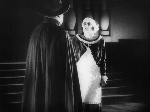 A black-and-white film still. A person in a dress looks in terror at another person, who wears a cape and top hat and is seen from behind. A staircase is visible in the background. 