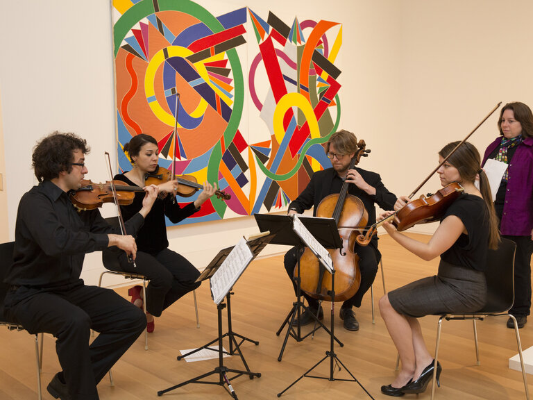 Four seated people playing string instruments in a gallery space. A colorful artwork hangs on the wall behind them. At right, a standing woman looks on. 