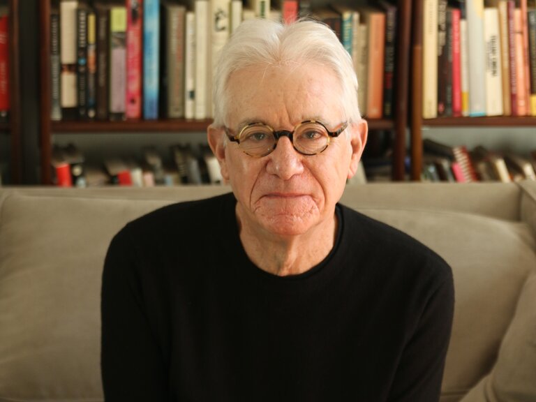 A seated man seen from the waist up. He looks into the camera with a gentle expression. He has short white hair and wears glasses with round, tortoise-shell frames, along with a black, long-sleeve top. 