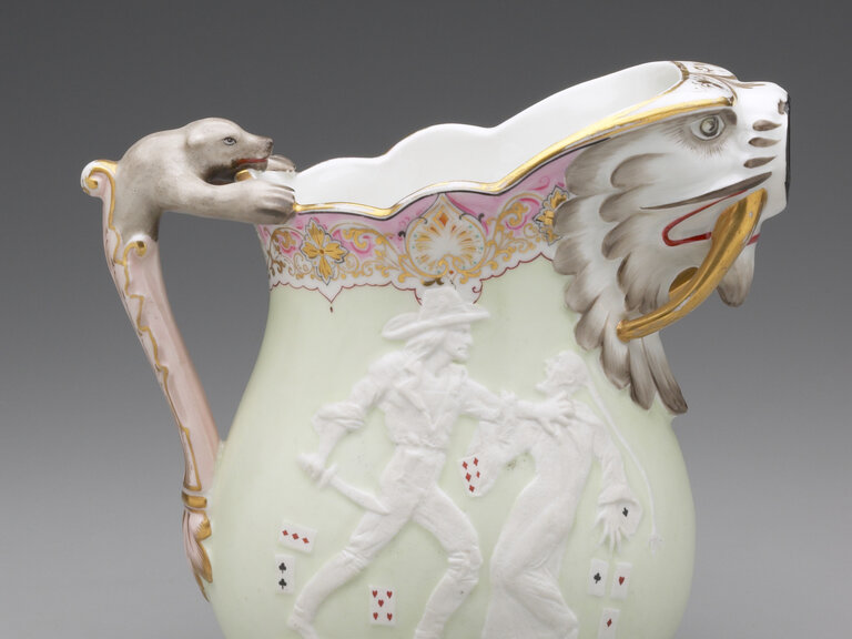 A pitcher seen in profile, with its handle at left and its spout at right. Both the handle and spout take the form of animals. On the body of the pitcher is a scene of a man grabbing another man with his left hand while wielding a knife with his right. Small playing cards surround them. 