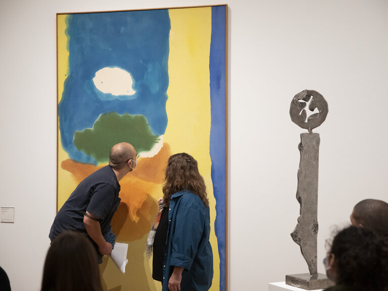 Two people seen from behind as they look closely at a large, abstract painting that hangs on the wall. To their right, an abstract sculpture stands on a platform. A few onlookers are seen from behind in the foreground.