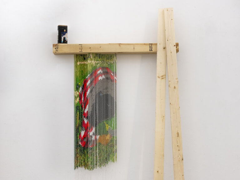 A horizontal piece of wood hangs on a wall. A small object stands on it, at far left. From this shelf are suspended strings of beads or fabric, which come together to form an abstract image. Two pieces of wood stand vertically, leaning against the horizontal piece’s right edge. 