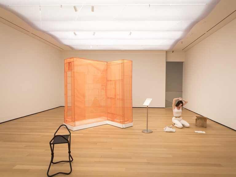 A person seated on their knees on the floor of a gallery. Next to them is a boxy orange object resembling a dresser but in an L shape