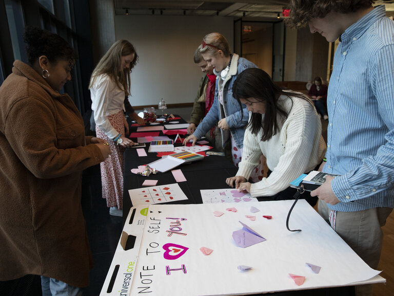 People stand gathered around a table covered in various papers. Some people focus on and handle items on the table. In the foreground, a large pad of paper reads NOTE TO SELF: I HEART YOU!