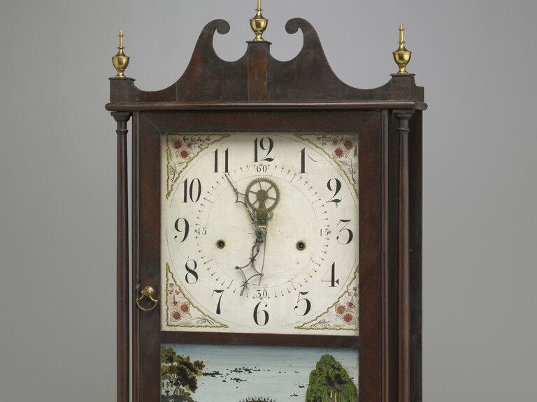 A clock in a wooden case. The case has four small, simple feet, while its top features three golden pieces resembling spires. The central spire is flanked by pieces of wood carved in a wavelike form. The clock appears above a scene with a tree at right and a building at left.