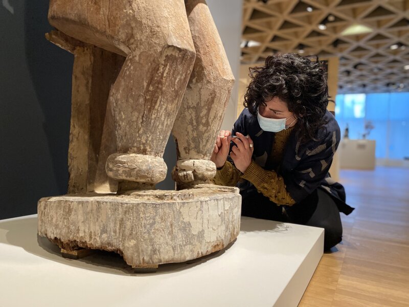 A woman with dark hair and a face mask kneels at the base of a wooden sculpture. She has an object in her hands and appears to be injecting the wooden sculpture with somethingd
