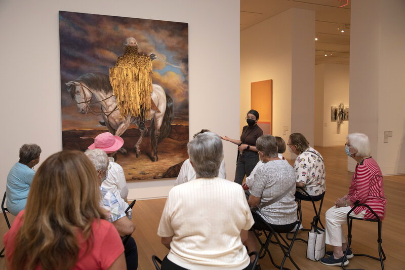 A group of older visitors sit facing a large painting of George Washington on a horse. George has strips of canvas nailed to his face and neck, covering his body. A teacher stands facing the visitors, gesturing towards the painting