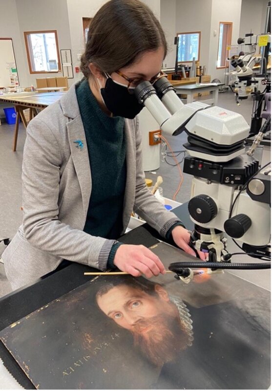 A conservator looks through a microscope while cleaning a portrait of a bearded man.