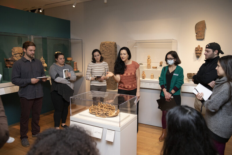 University students gather around a ceramic object on a pedestal. There are additional art objects on the walls and in cases behind the students